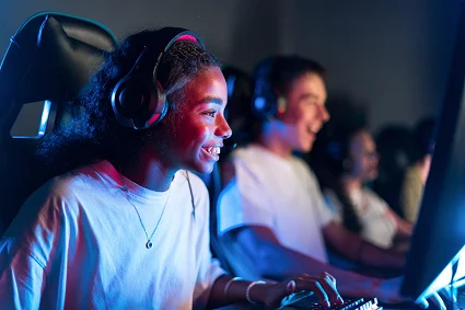 Een vrouwelijke gamer met een headset op, die glimlachend op een computer speelt terwijl haar teamgenoten op de achtergrond te zien zijn; ze geniet van een online multiplayer-spelsessie